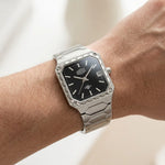 A silver-toned metal wristwatch with a black dial and date window, shown worn on a person's wrist against a soft light background.