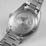 Close-up of the back of a stainless steel wristwatch with a metal link band, showing an engraved desert scene with palm trees, a camel, and pyramids. The watch case and band are silver-toned metal on a light gray background.