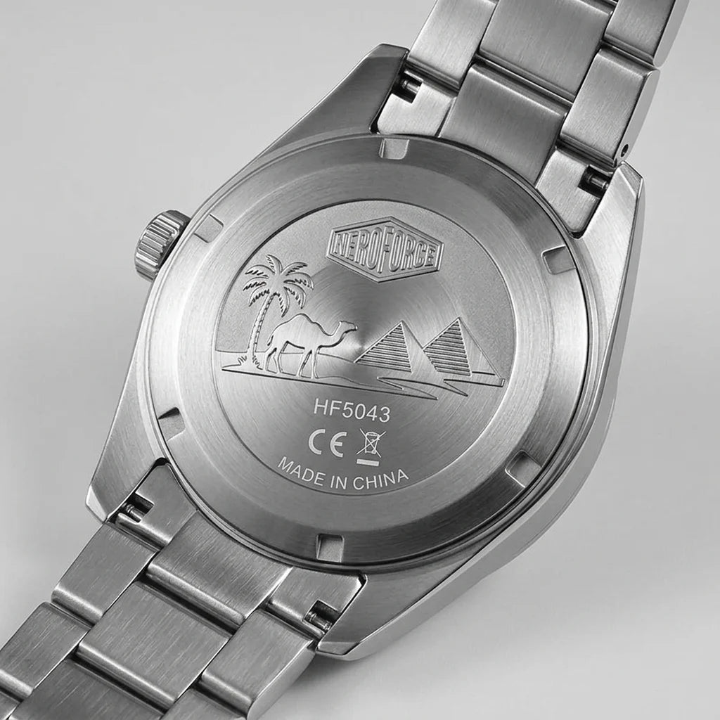 Close-up of the back of a stainless steel wristwatch with a metal link band, showing an engraved desert scene with palm trees, a camel, and pyramids. The watch case and band are silver-toned metal on a light gray background.