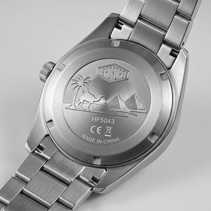 Close-up of the back of a stainless steel wristwatch with a metal link band, showing an engraved desert scene with palm trees, a camel, and pyramids. The watch case and band are silver-toned metal on a light gray background.